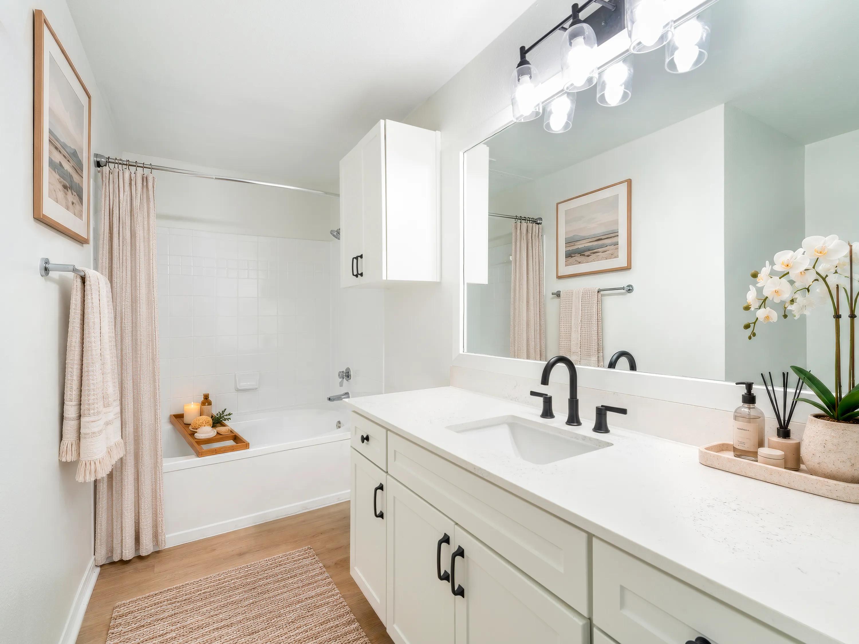 Light finish bathroom with white shaker-style cabinetry, quartz countertops, black fixtures, a large vanity mirror, wood-style flooring, and a soaking tub at Alister Sunset Valley apartments in Austin, TX.