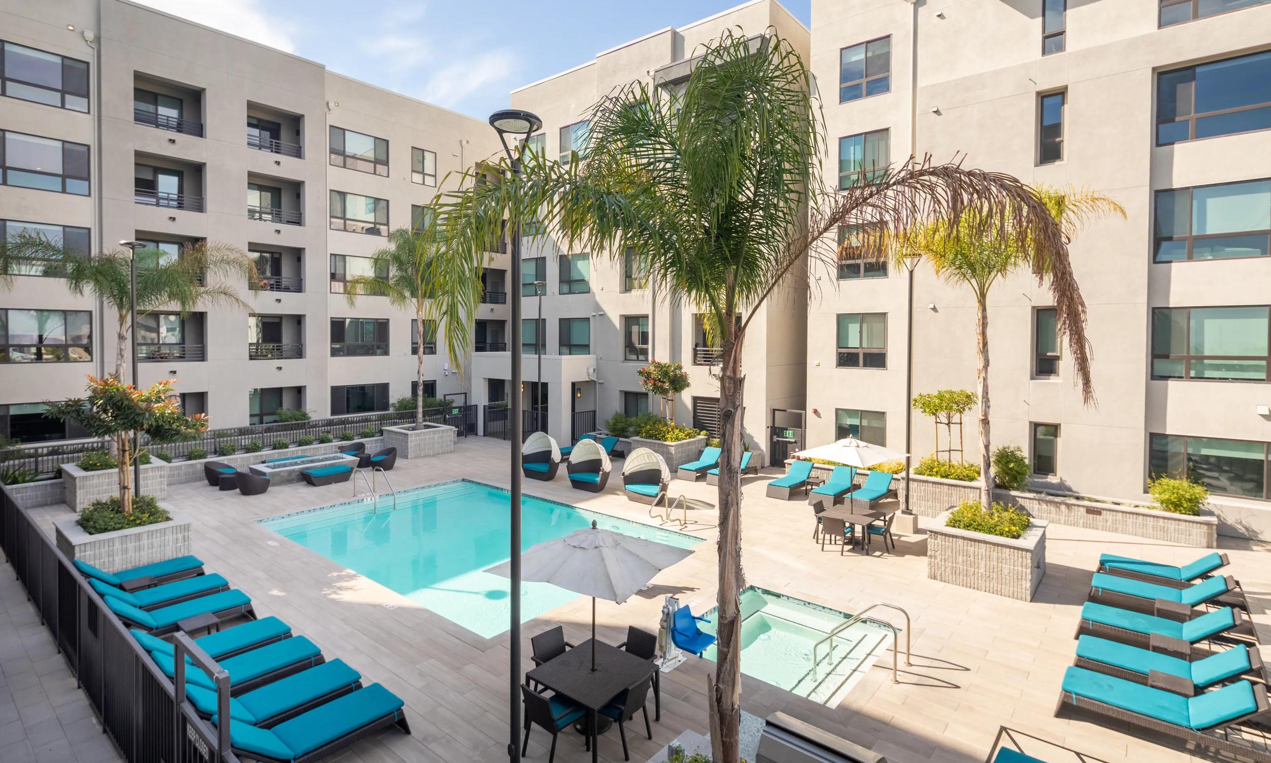 Luxurious apartment pool area with modern lounge chairs, lush palm trees, and scenic city views.