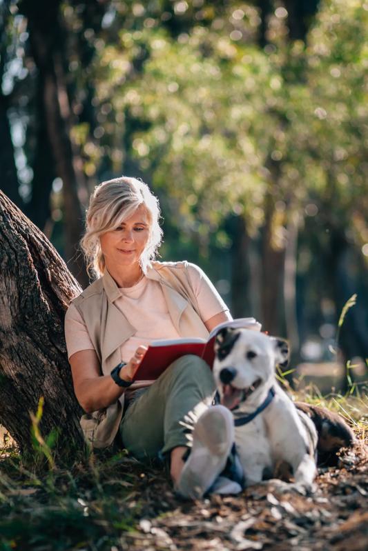 Mature woman reading outside under tree with dog near Modera Natick Center apartments for rent
