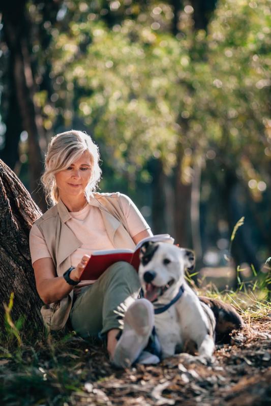 mature woman with dog reading a book leaded up against a tree in a park alister oak hill apartments in austin tx