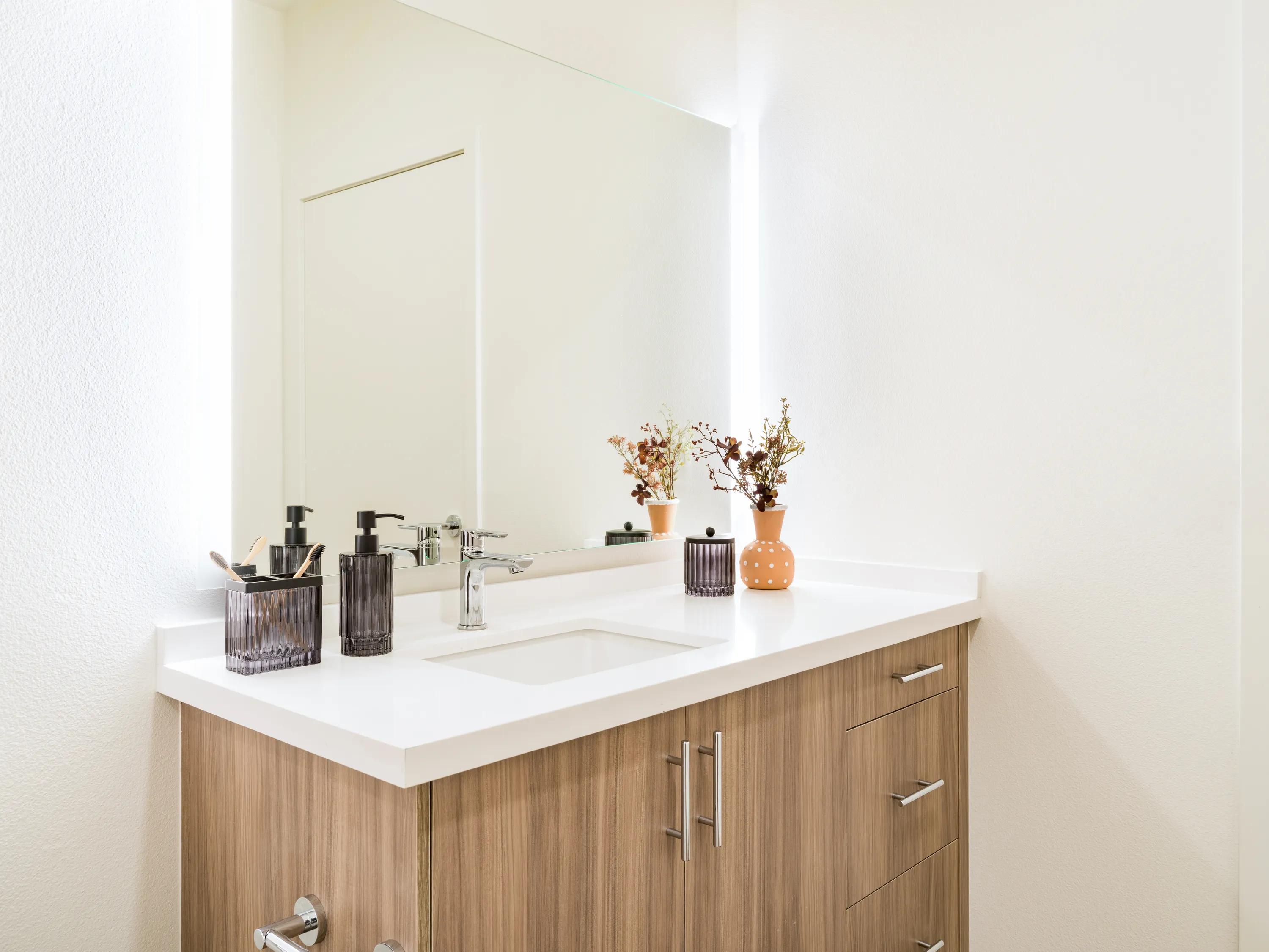 Contemporary bathroom vanity with a quartz-style countertop, wood-grain cabinetry, integrated sink, modern fixtures, and an oversized mirror, showcasing one of several available interior finish palettes at Modera Melrose apartments for rent in Oceanside, CA.