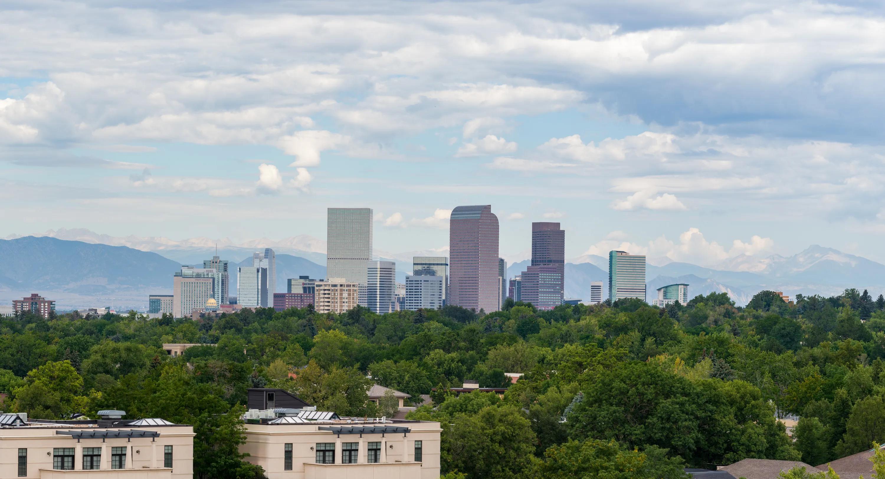 View of the Denver skyline and surrounding mountain range from an elevated vantage point, overlooking a tree-lined neighborhood near Modera Second Avenue apartments in Denver, CO.