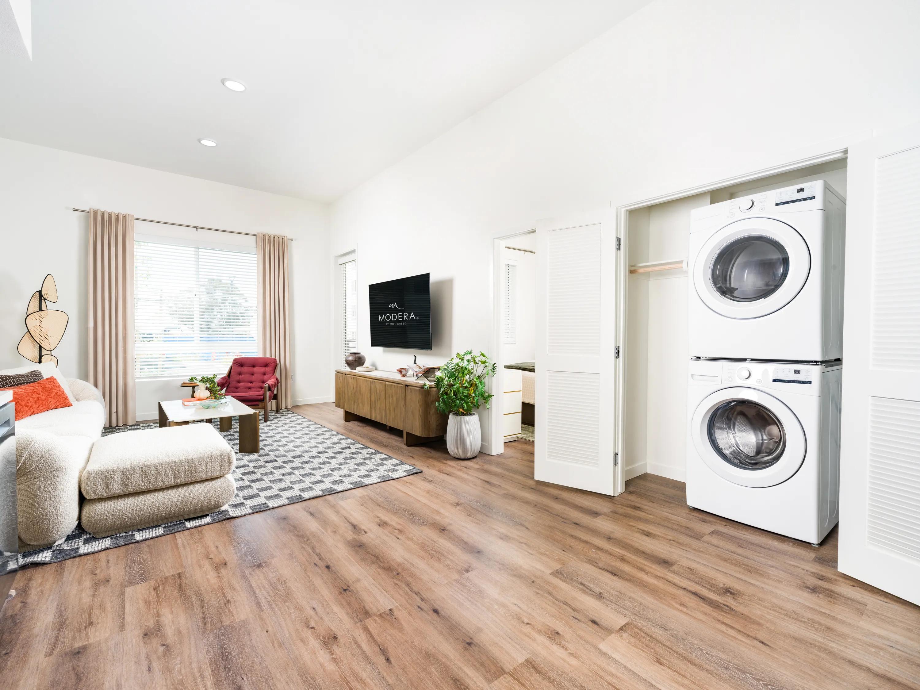 Open-concept living room with wood-style flooring, high ceilings, an in-home stacked washer and dryer, and contemporary furnishings at Modera Melrose apartments for rent in Oceanside, CA.