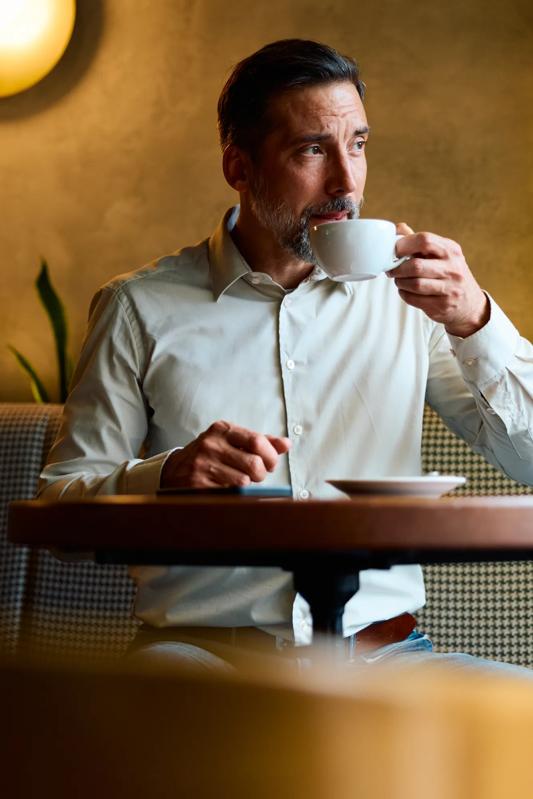 Mature bearded man in smart casual attire sits alone in a modern cafe, sipping coffee and gazing away, deep in thought about business plans, strategy and future possibilities modera second avenue apartments in denver co