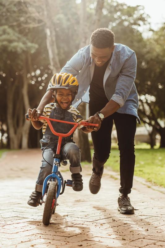 dad teaching young son to ride a bike while holding the bicycle both smiling and happy outdoors in a park denver co