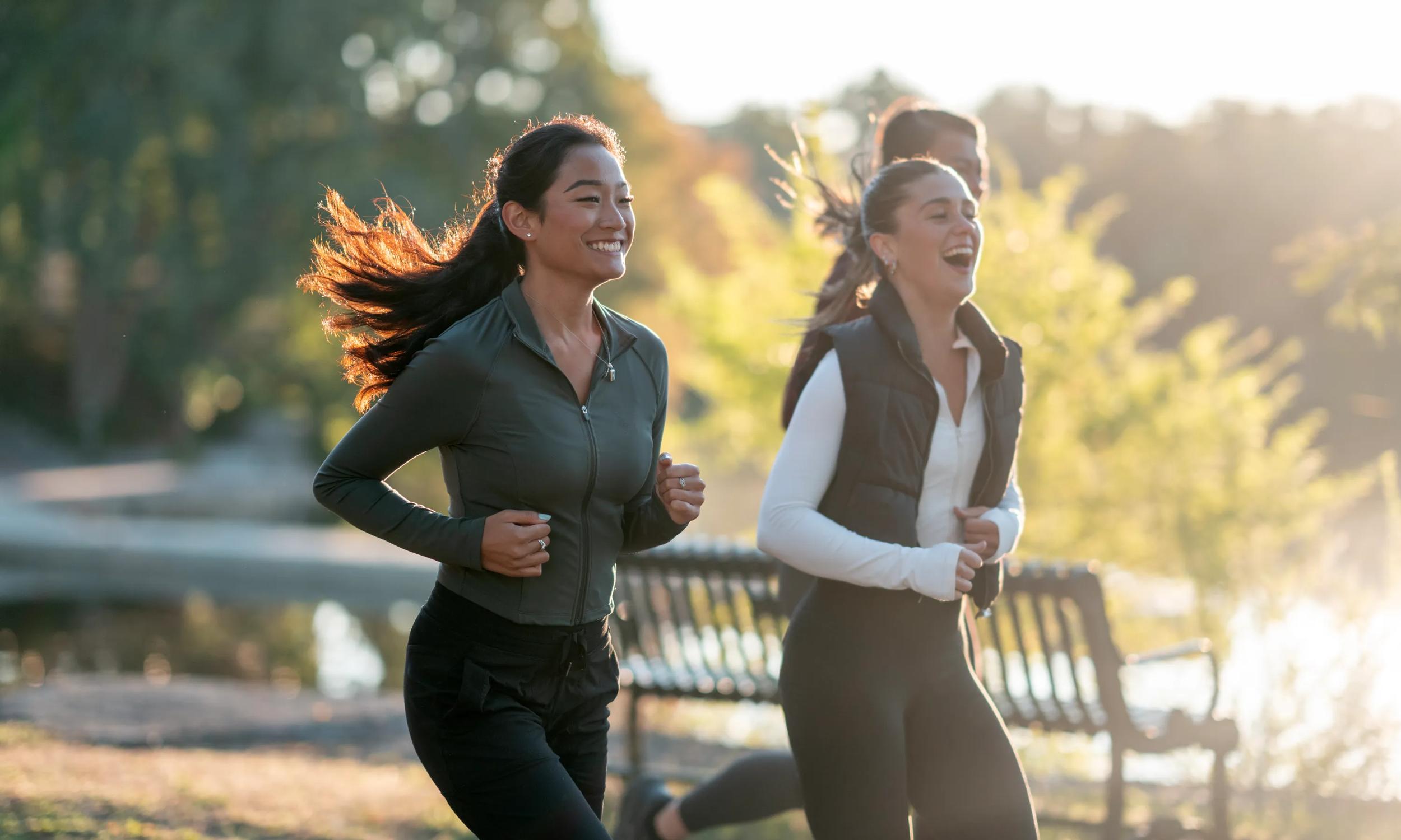 Two joyful women are jogging in a park on a sunny day, showcasing active lifestyles. Sunlight filters through the trees, enhancing the vibrant mood and portraying fitness and togetherness during outdoor activities modera second avenue apart