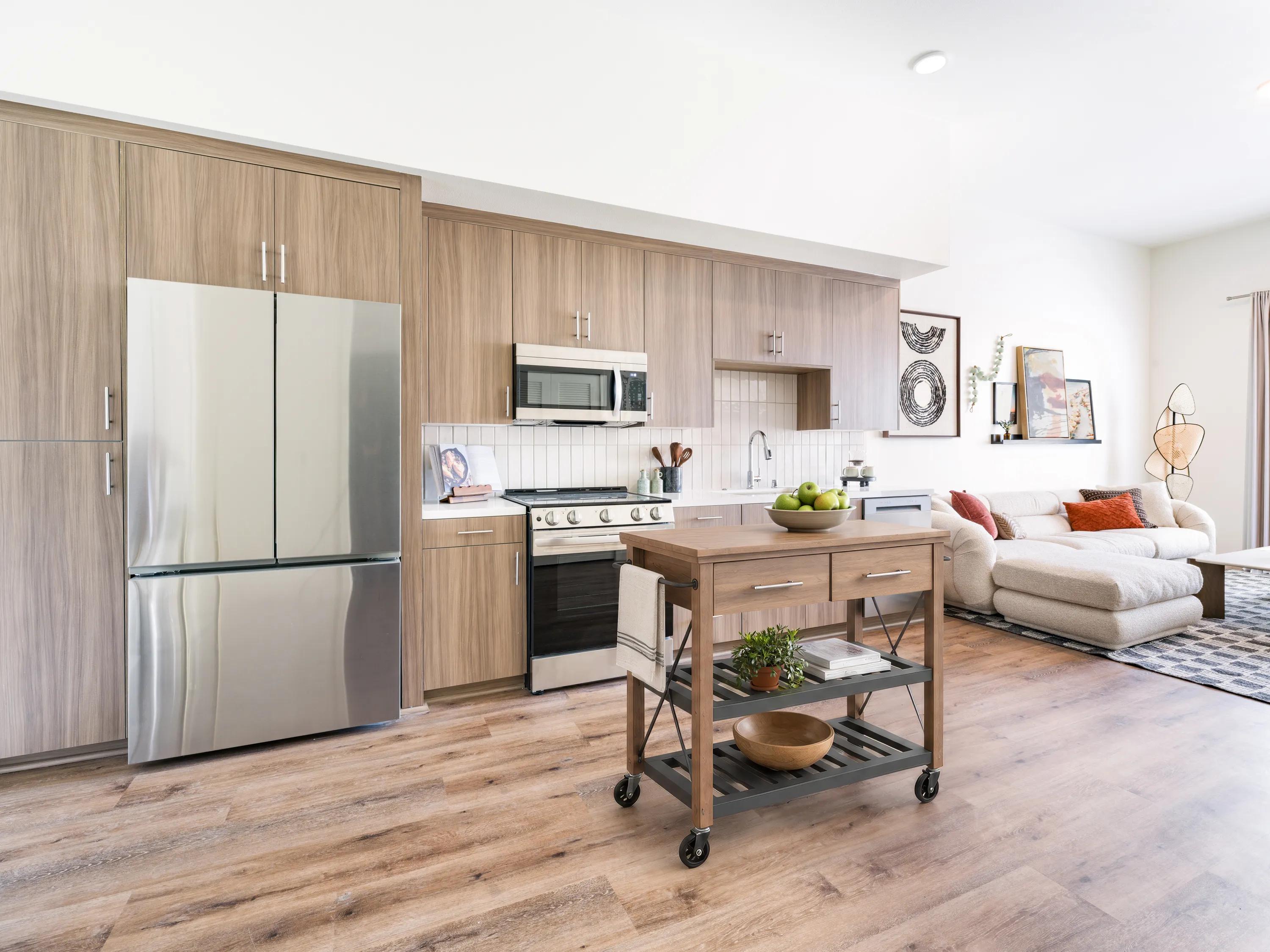 Modern kitchen with stainless steel appliances, wood-look cabinetry, a movable kitchen island, and open-concept living space at Modera Melrose apartments for rent in Oceanside, CA.