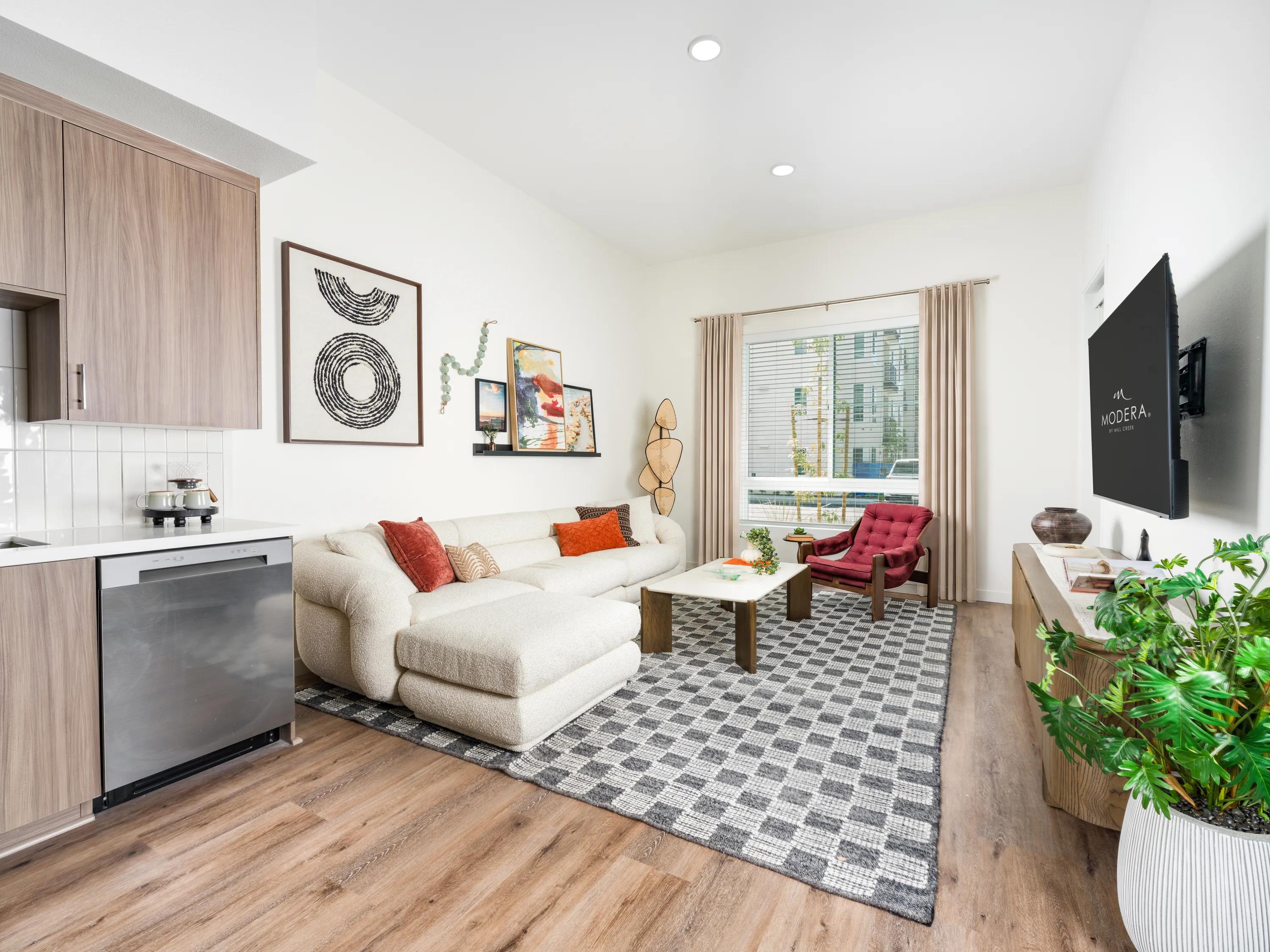 Open-concept living room with contemporary furnishings, wood-style flooring, oversized windows, and seamless access to the kitchen at Modera Melrose apartments for rent in Oceanside, CA.