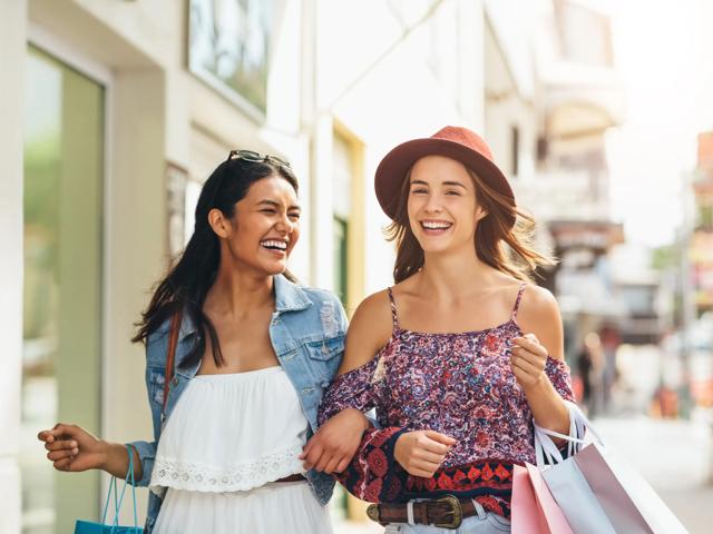two young women holding shopping bags at outdoor mall smiling and linked arm in arm alister sherman oaks