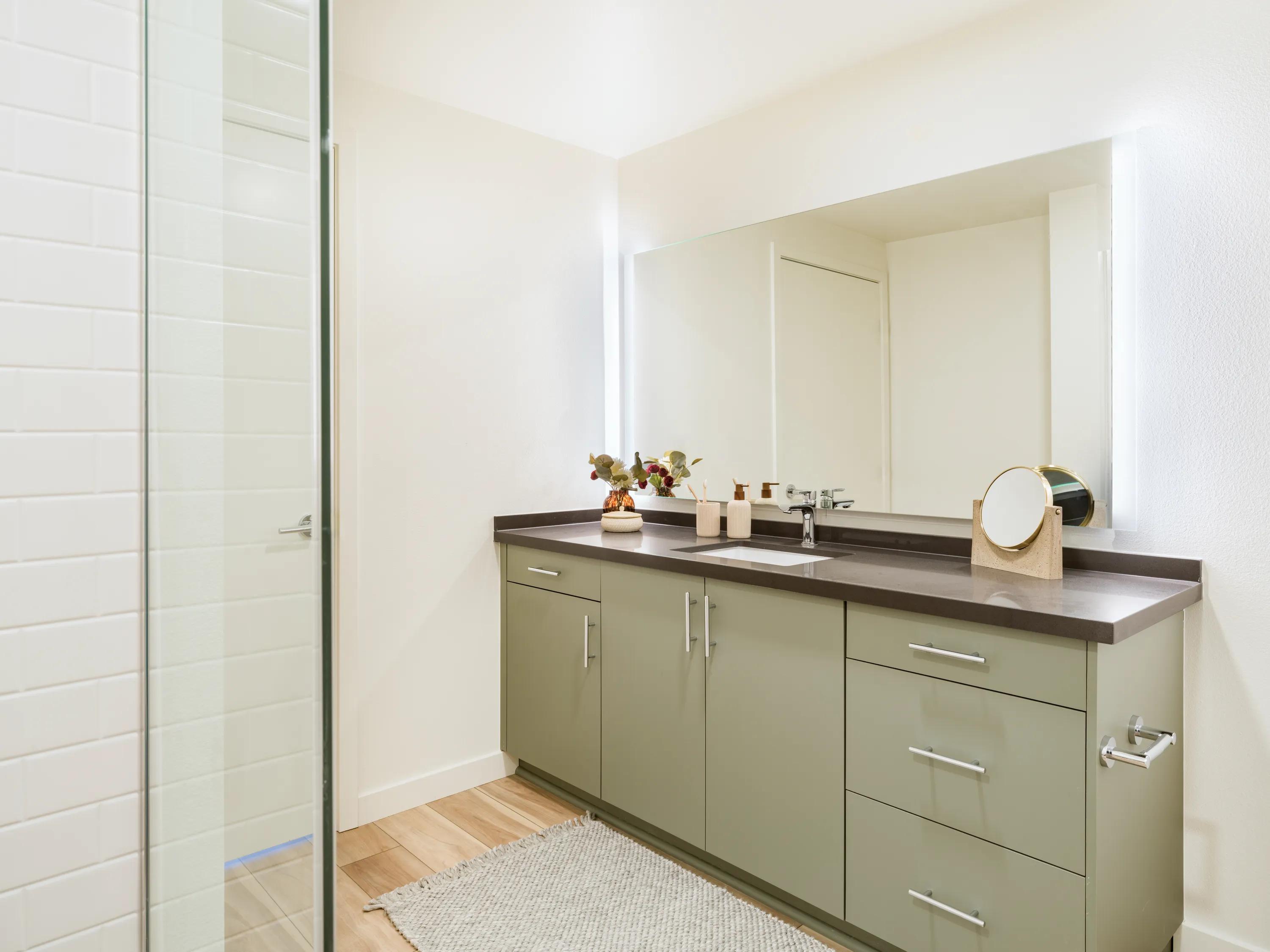 Contemporary bathroom with a spacious vanity, quartz-style countertop, modern cabinetry, oversized mirror, wood-style flooring, and a frameless glass shower at Modera Melrose apartments for rent in Oceanside, CA.