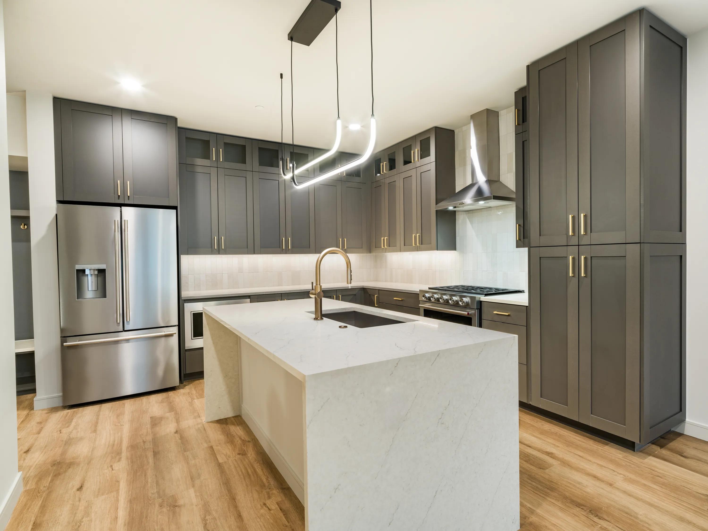 Gourmet kitchen with a quartz waterfall island, full-height gray cabinetry, stainless steel appliances, wood-style flooring, and modern pendant lighting at Modera Second Avenue apartments in Denver, CO.